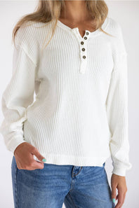 A woman wearing a white waffle-knit long-sleeve henley sweater with three dark buttons, paired with blue jeans, against a plain white background.