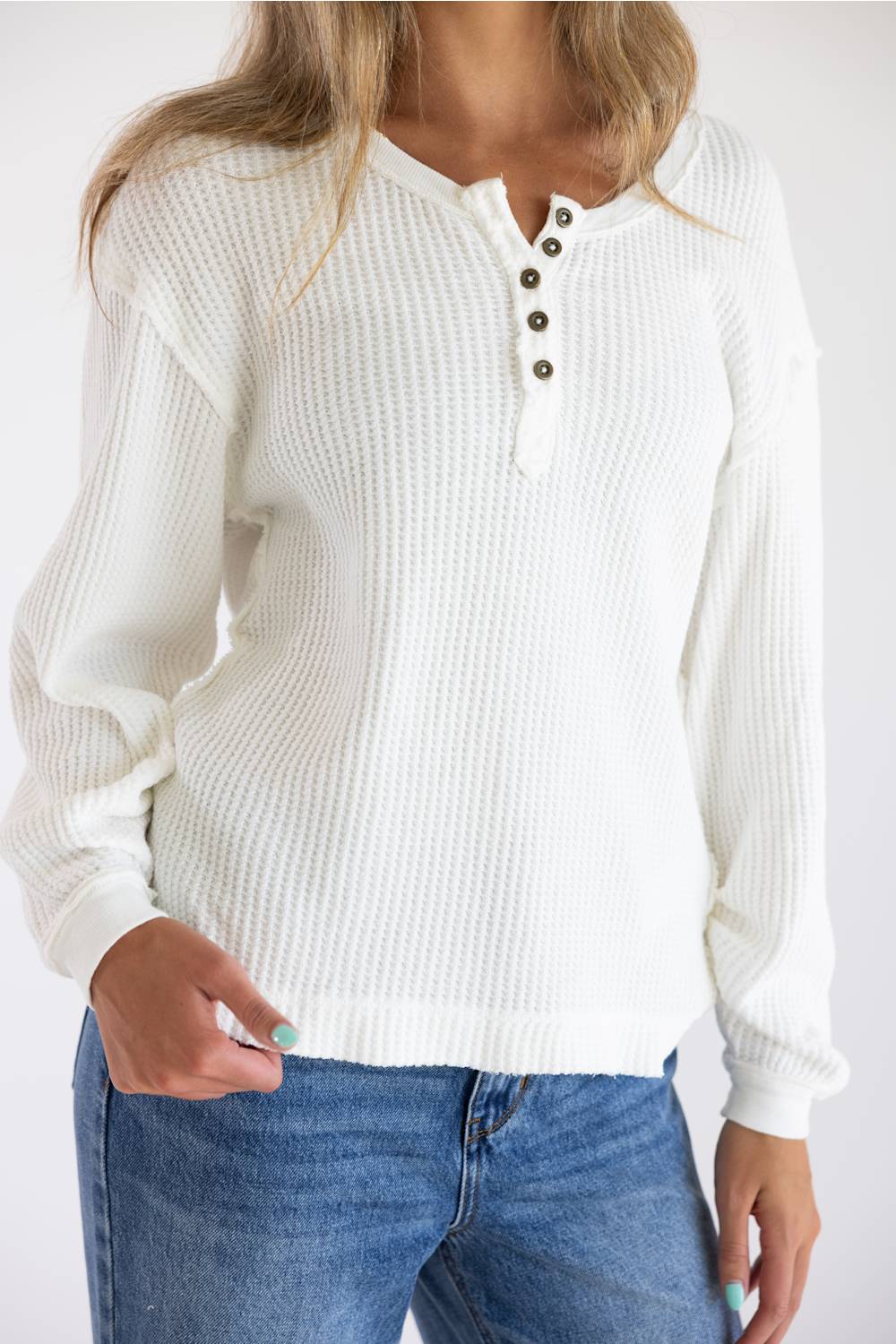 A woman wearing a white waffle-knit long-sleeve henley sweater with three dark buttons, paired with blue jeans, against a plain white background.
