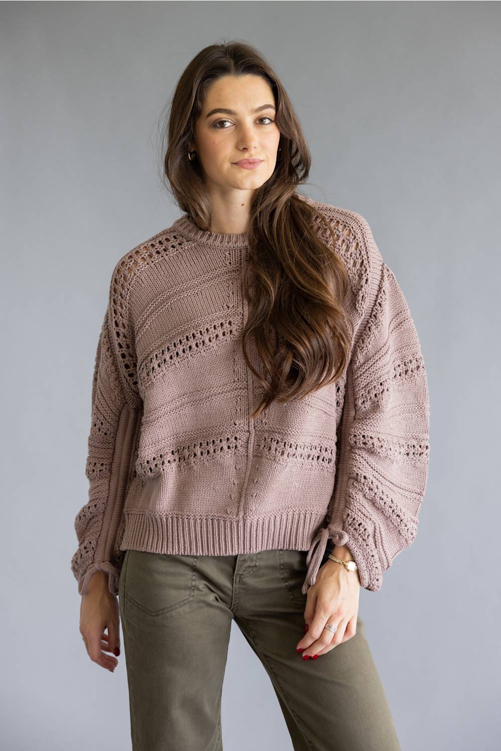 A woman modeling a dusty rose knit sweater with textured horizontal patterns and side tie details, paired with olive green pants, against a solid gray studio background.
