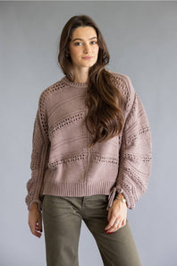 A woman modeling a dusty rose knit sweater with textured horizontal patterns and side tie details, paired with olive green pants, against a solid gray studio background.