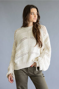 A woman wearing a cream-colored, open-knit cropped sweater with tie details on the cuffs, paired with olive green pants, set against a solid gray studio background.