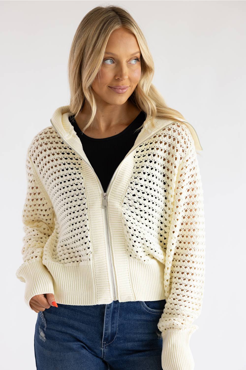 A woman wearing a cream-colored open-knit zip-up hoodie with a ribbed hem and cuffs, layered over a black top and paired with blue jeans, against a plain light gray background.