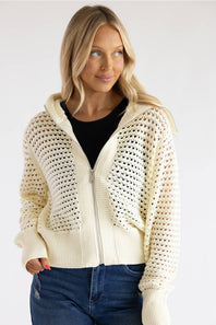 A woman wearing a cream-colored open-knit zip-up hoodie with a ribbed hem and cuffs, layered over a black top and paired with blue jeans, against a plain light gray background.
