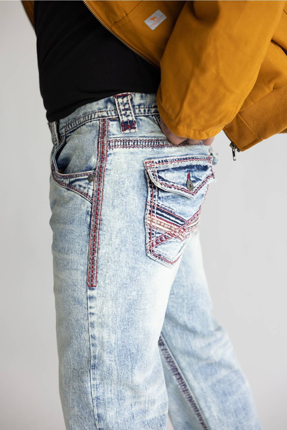 Close-up of a person wearing light-wash blue jeans with decorative red and dark contrast stitching, paired with a black top and mustard yellow jacket against a plain light gray background.