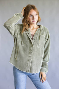 A woman modeling an oversized sage green corduroy button-up shirt jacket with front chest pockets, paired with light blue jeans, against a neutral gray studio background.