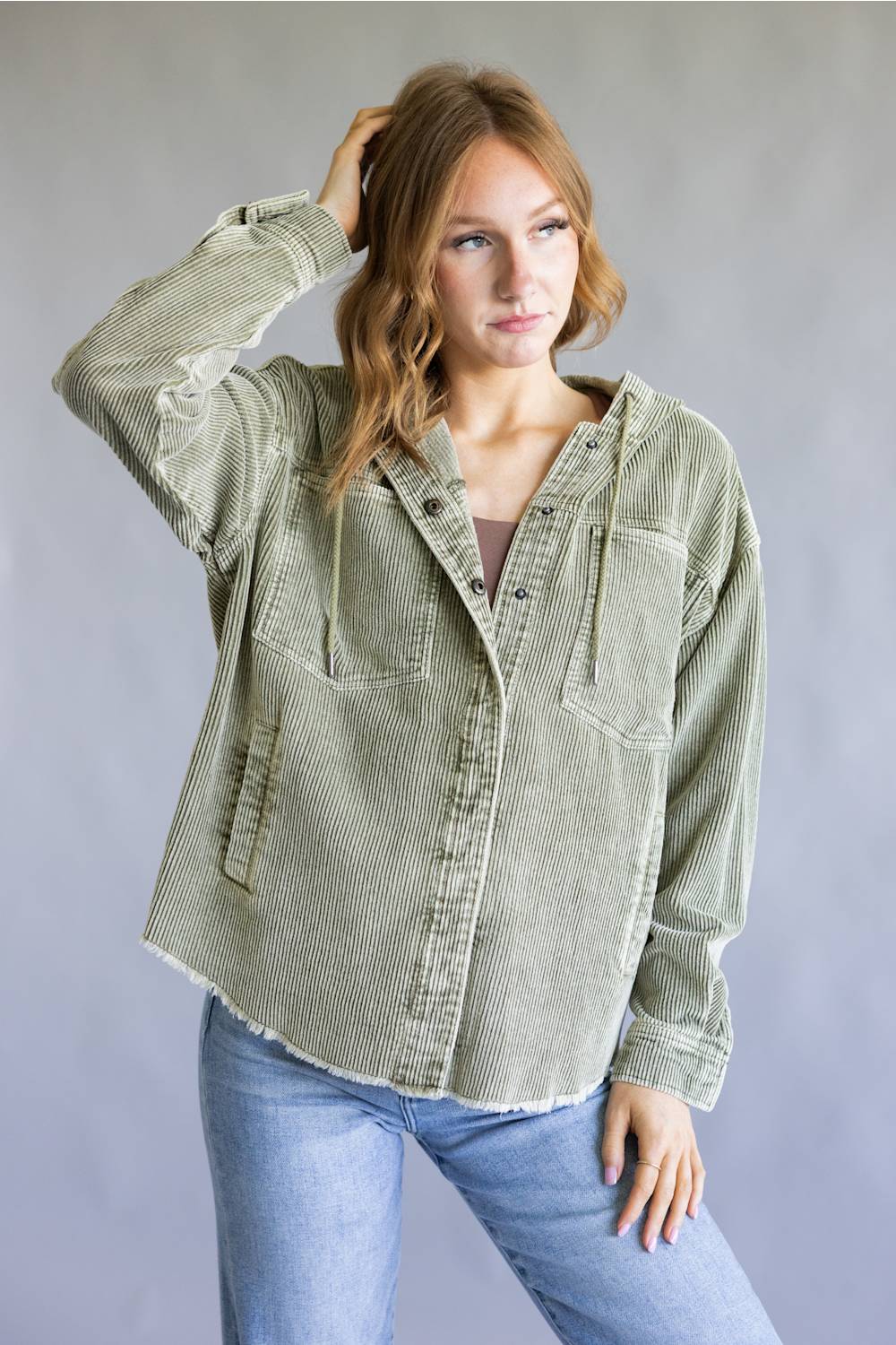 A woman modeling an oversized sage green corduroy button-up shirt jacket with front chest pockets, paired with light blue jeans, against a neutral gray studio background.