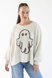 A woman wearing a cream-colored knit sweater with a black and light orange gingham ghost graphic on the front, paired with blue jeans, against a plain white background.