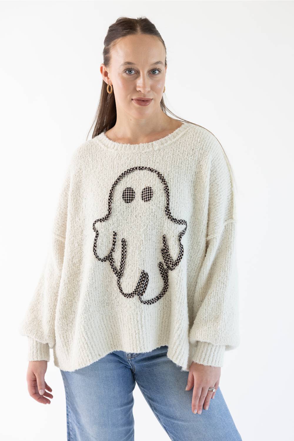 A woman wearing a cream-colored knit sweater with a black and light orange gingham ghost graphic on the front, paired with blue jeans, against a plain white background.