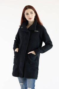 A woman with red hair wearing a black sherpa fleece coat with side pockets and a small white embroidered logo on the chest, paired with blue jeans, against a plain white background.