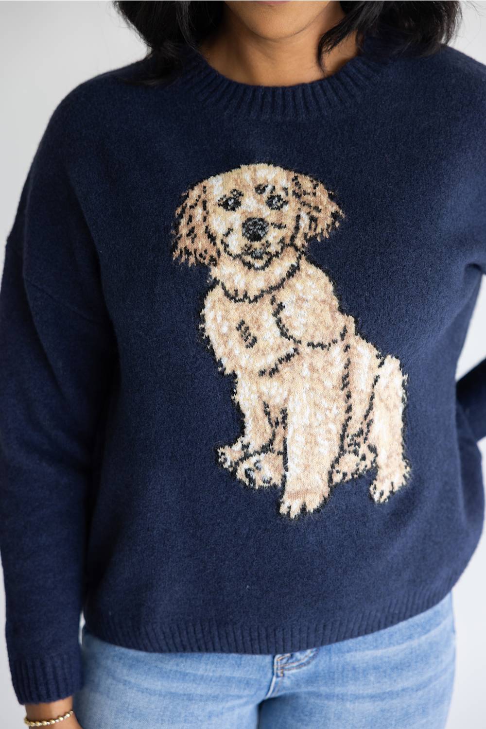 A person wearing a navy blue knit sweater with a large beige and cream dog graphic on the front, paired with light blue jeans.