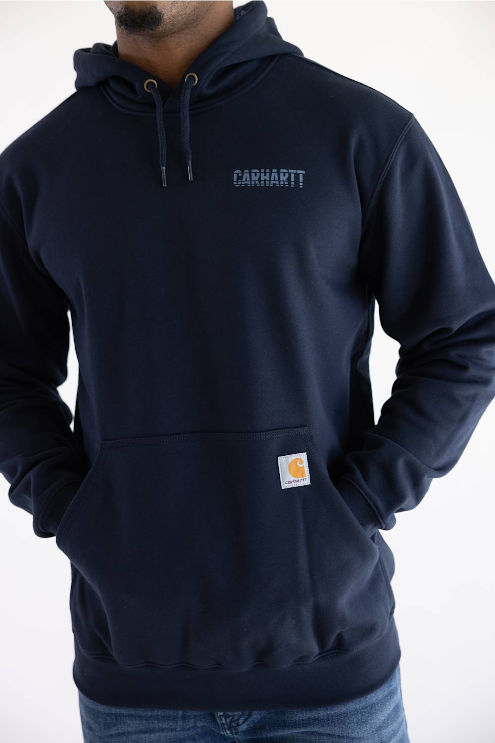 A man wearing a navy blue Carhartt pullover hoodie with a small embroidered Carhartt logo on the left chest and a signature yellow-and-white brand patch on the front pocket, paired with blue jeans against a plain white background.