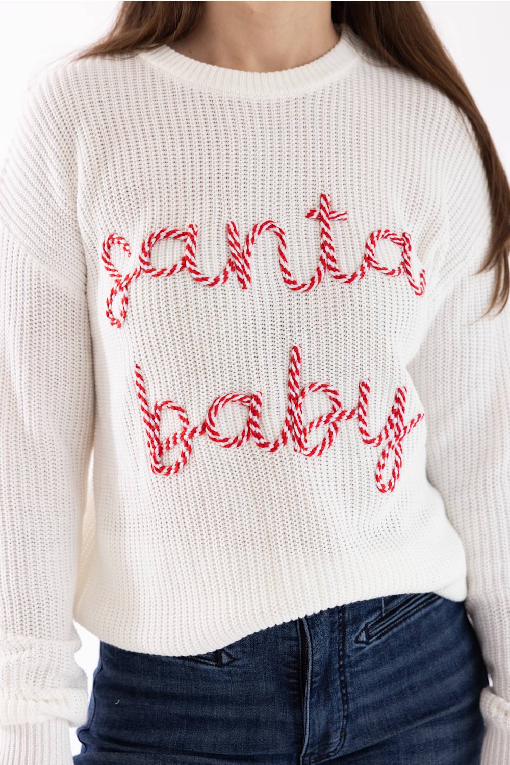 A person wearing a white ribbed knit sweater with 'santa baby' written across the front in red and white candy cane lettering, paired with dark blue jeans.