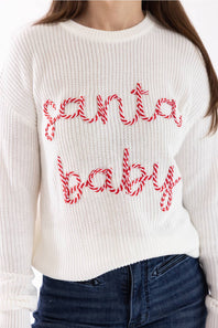 A person wearing a white ribbed knit sweater with 'santa baby' written across the front in red and white candy cane lettering, paired with dark blue jeans.