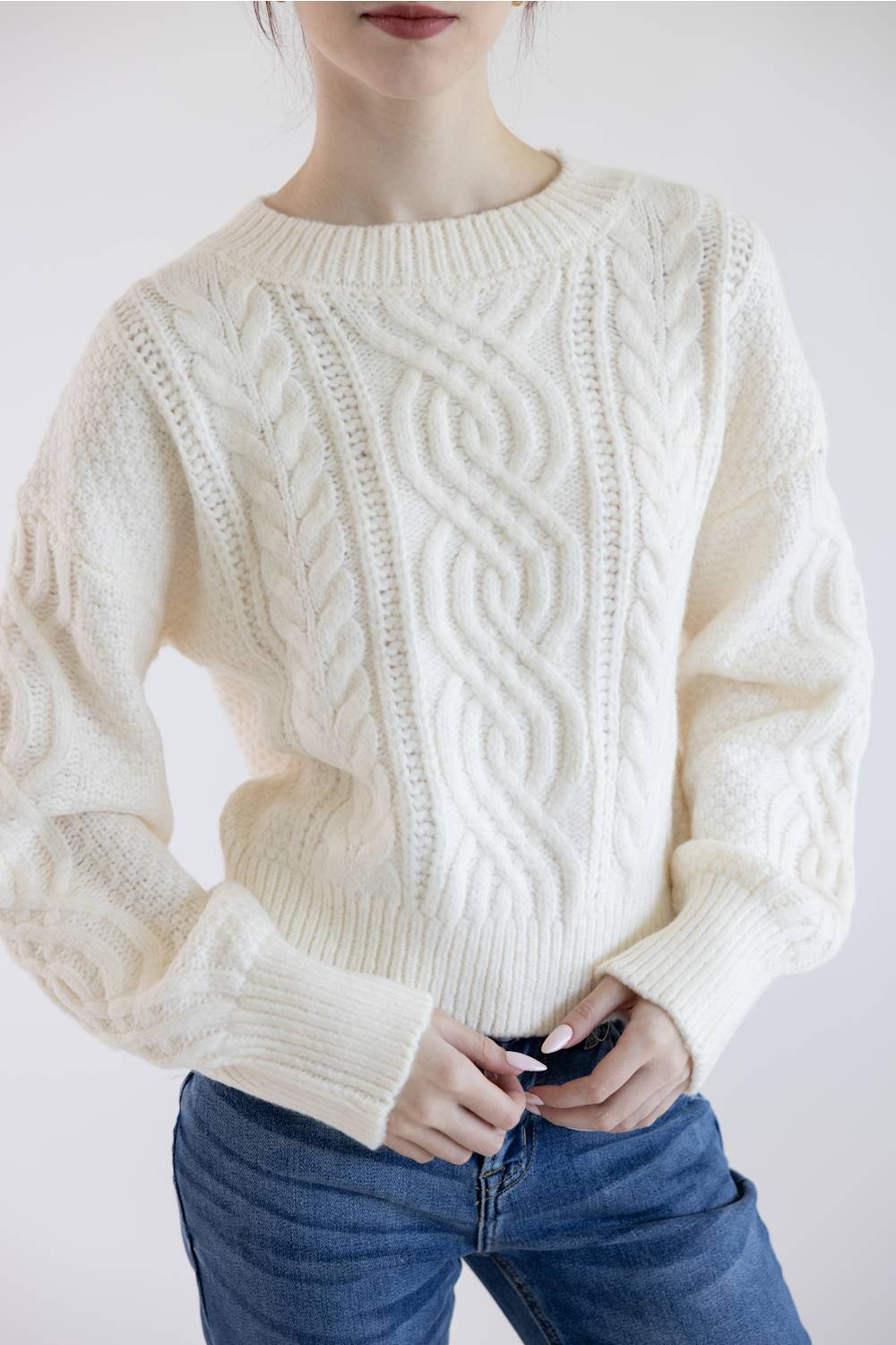 A woman wearing a cream-colored cable-knit sweater and blue jeans, shown from the chin down against a plain light background.