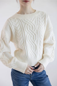 A woman wearing a cream-colored cable-knit sweater and blue jeans, shown from the chin down against a plain light background.