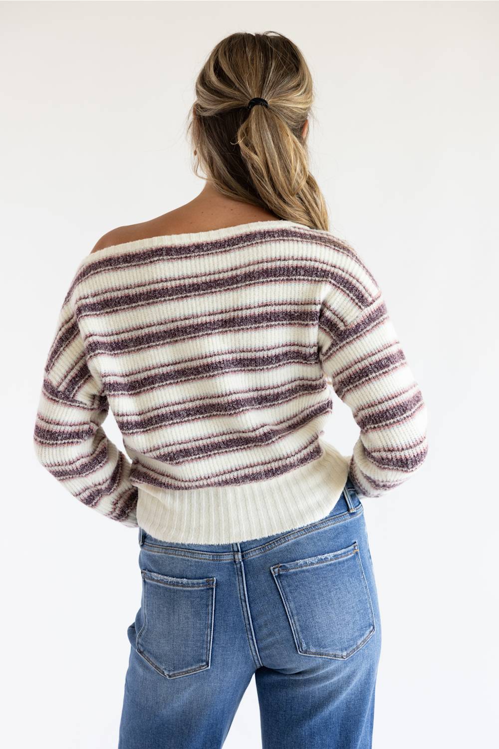 A woman shown from the back wearing a cream knit sweater with dark brown horizontal stripes and blue jeans, set against a plain white studio background.