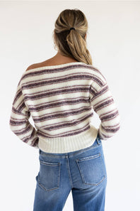 A woman shown from the back wearing a cream knit sweater with dark brown horizontal stripes and blue jeans, set against a plain white studio background.