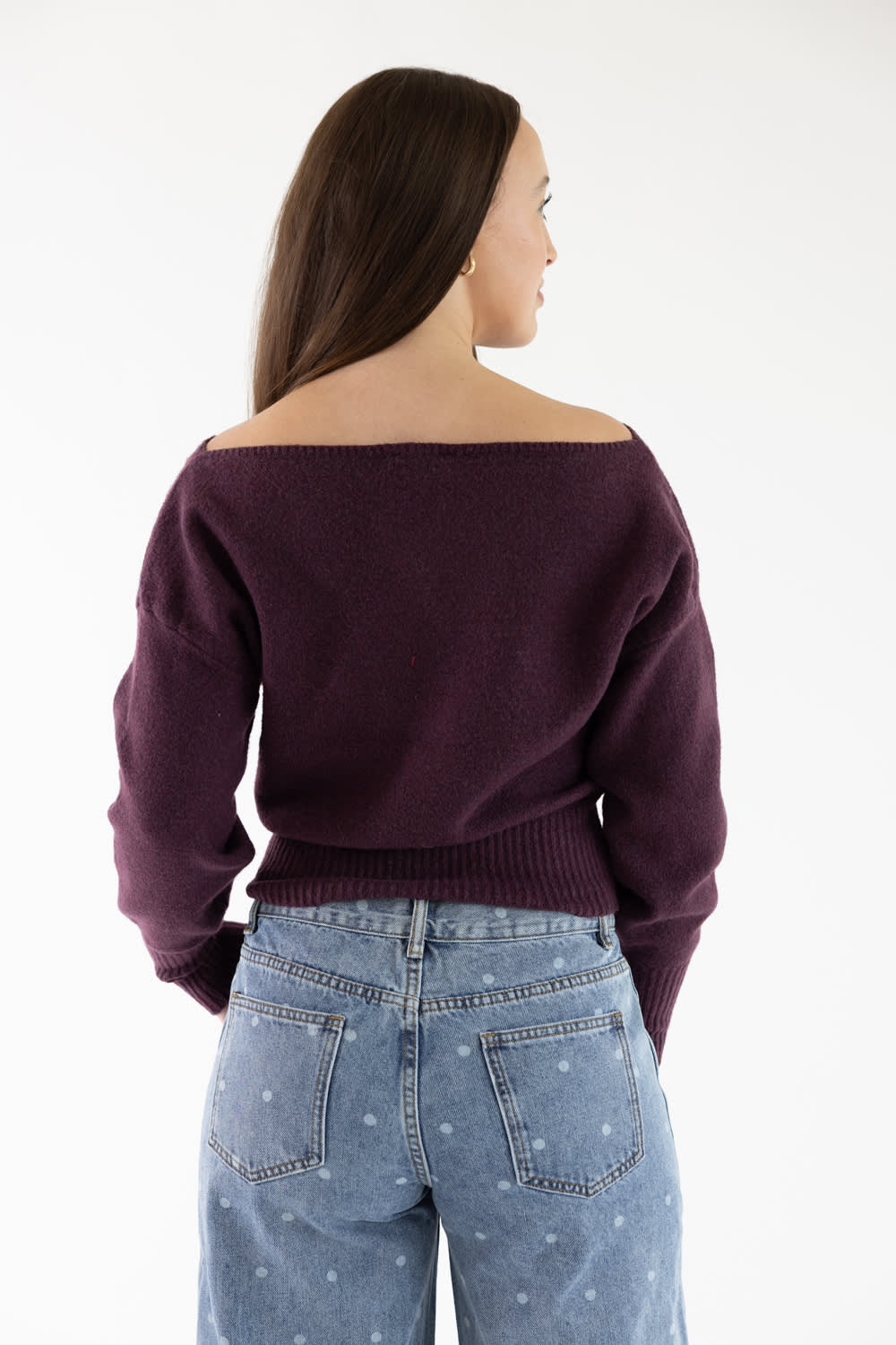 A woman shown from the back wearing a dark plum off-the-shoulder knit sweater and light blue jeans with small white dots, against a plain white studio background.