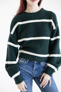A woman wearing a dark forest green knit sweater with horizontal cream-white stripes and blue jeans, shown from the neck down against a plain white background.