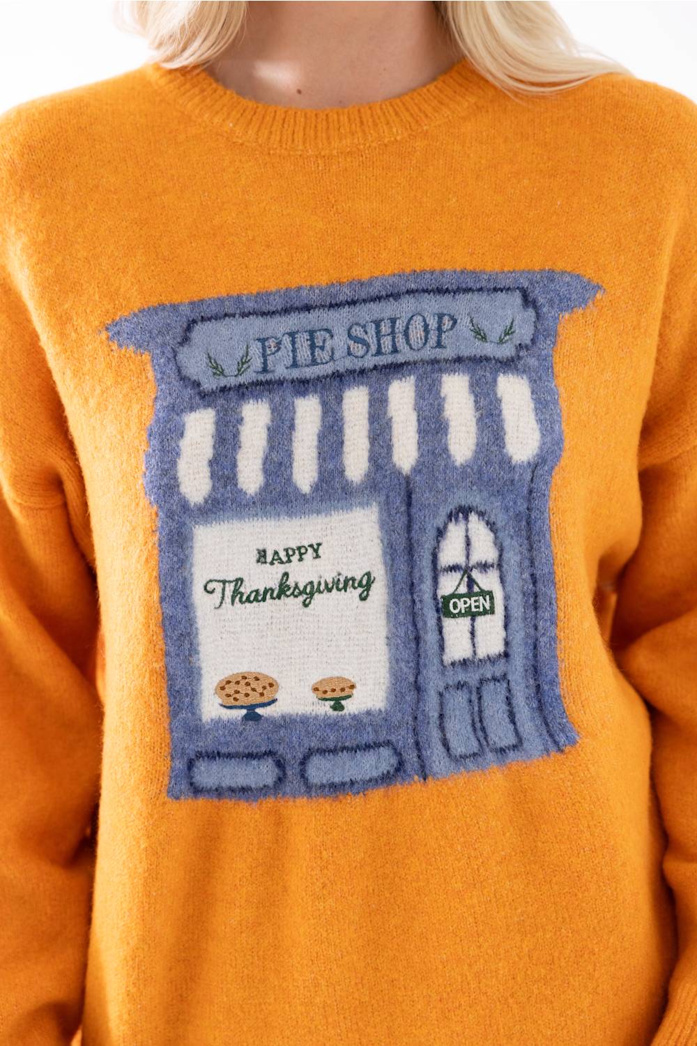 A close-up of a person wearing a bright orange knit sweater with a large blue pie shop graphic on the front that reads 'Happy Thanksgiving' and 'Pie Shop.'.