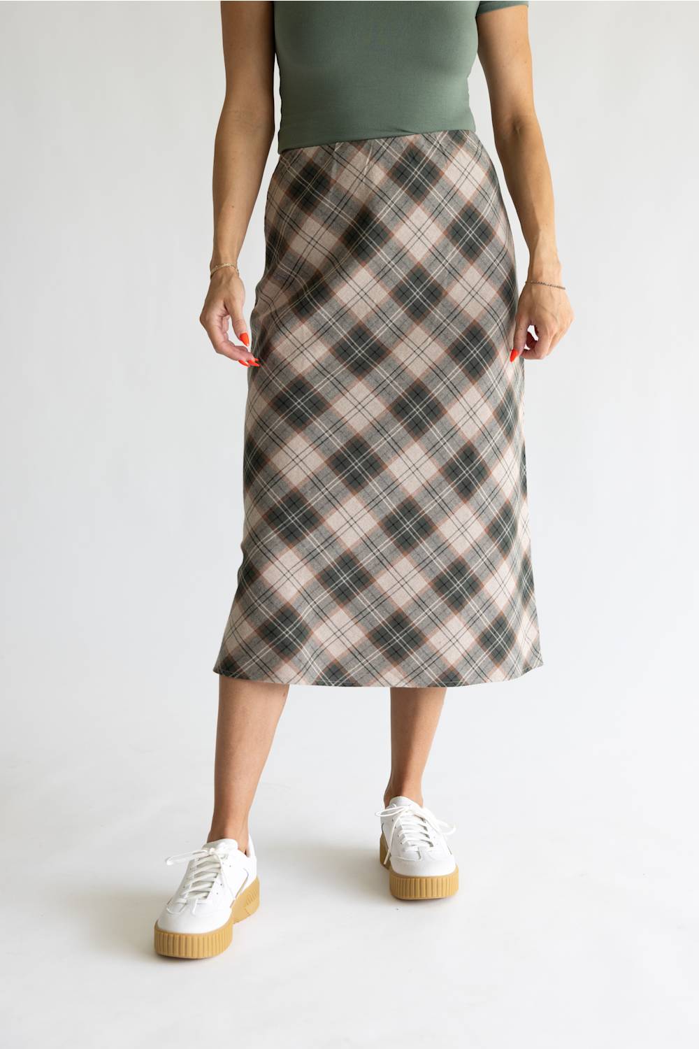 A model wearing a knee-length plaid midi skirt in beige, brown, and black, paired with a sage green top and white platform sneakers, against a plain light gray background.