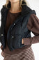 A close-up of a person wearing a black puffer vest over a brown ribbed long-sleeve top and a brown-and-white striped skirt, against a plain light background.