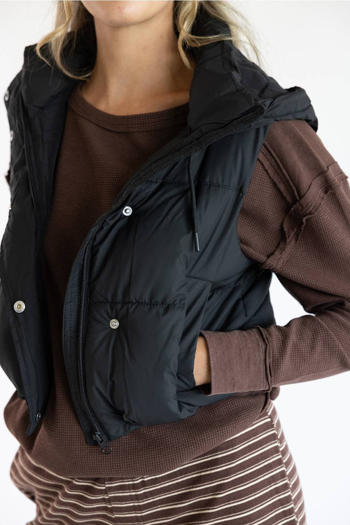 A close-up of a person wearing a black puffer vest over a brown ribbed long-sleeve top and a brown-and-white striped skirt, against a plain light background.