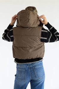 A person viewed from the back wearing a taupe-brown puffer vest over a black and white striped sweater and blue jeans, adjusting the hood of the vest against a plain white background.