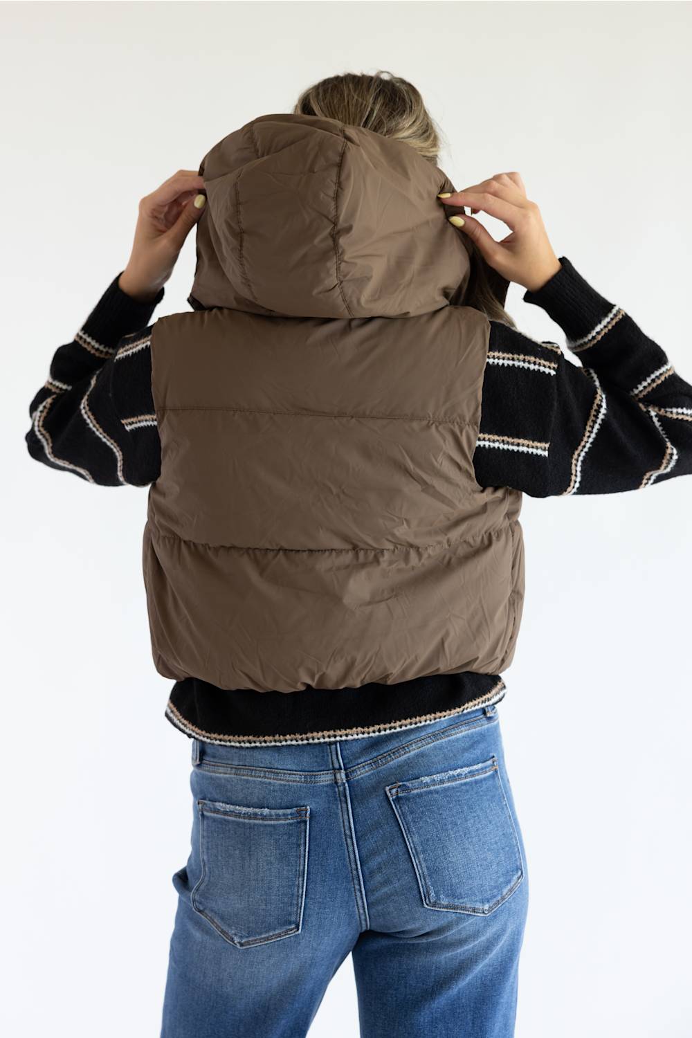 A person viewed from the back wearing a taupe-brown puffer vest over a black and white striped sweater and blue jeans, adjusting the hood of the vest against a plain white background.