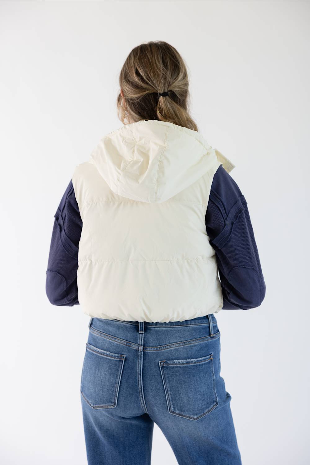 A woman viewed from behind wearing a cream hooded puffer vest over a navy blue long-sleeve top and blue jeans, against a plain white studio background.