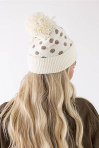A cream-colored knit beanie with brown polka dots and a matching cream pom-pom, shown on a model from behind against a plain white background.