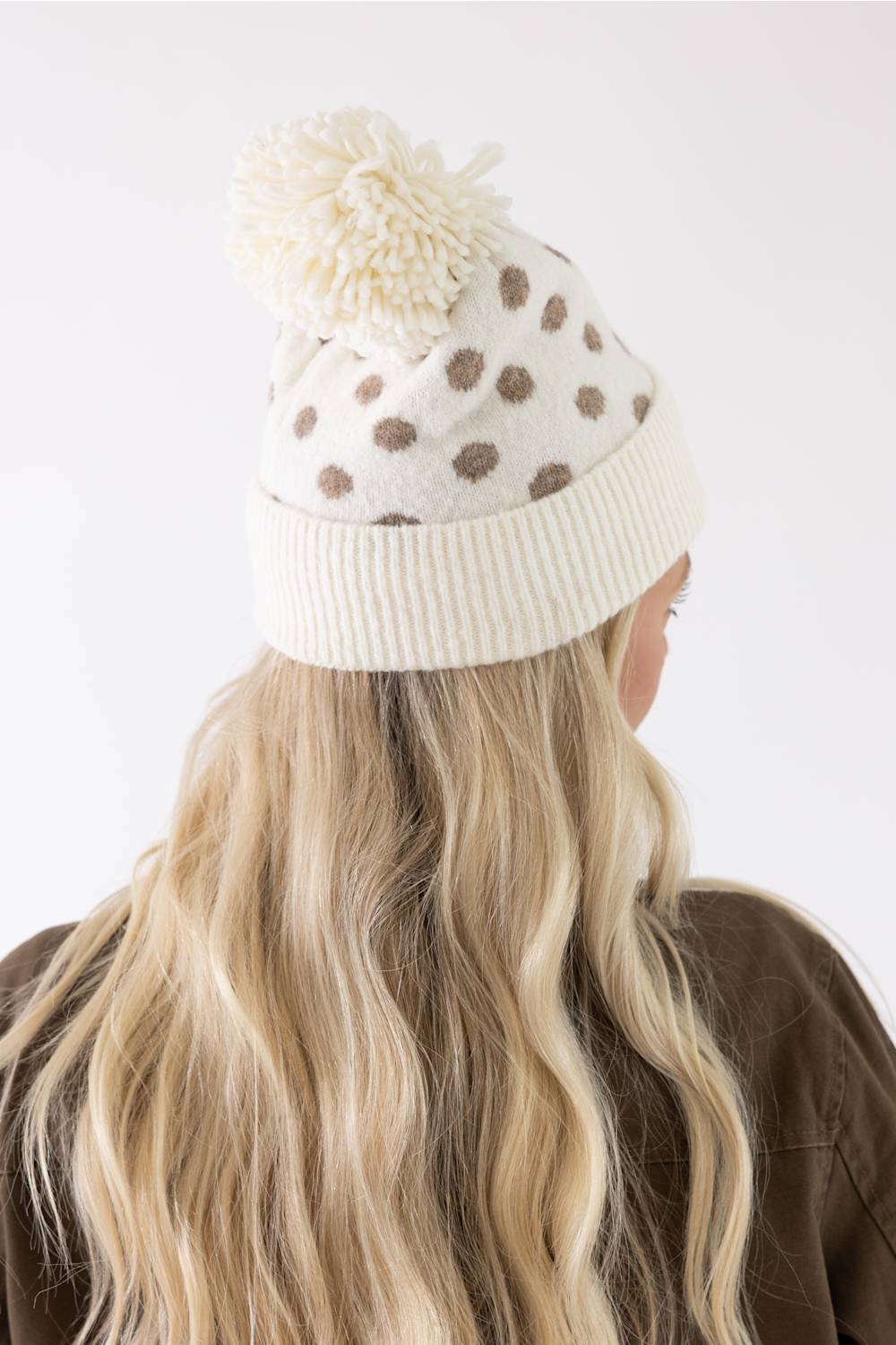 A cream-colored knit beanie with brown polka dots and a matching cream pom-pom, shown on a model from behind against a plain white background.