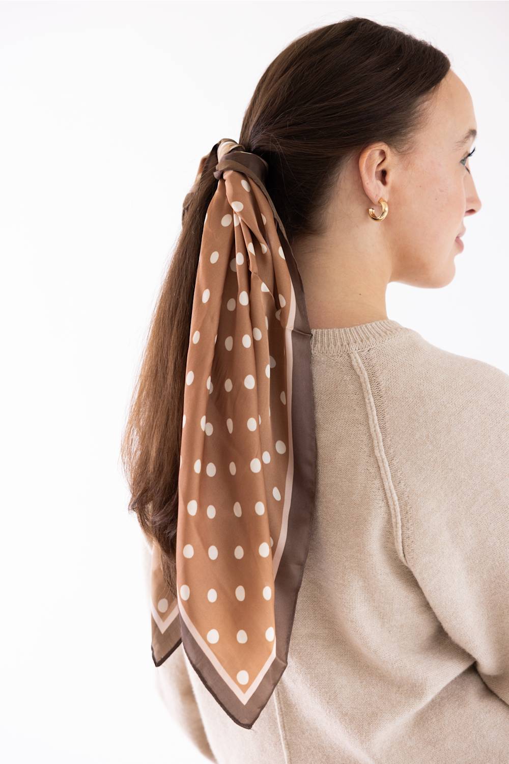 A woman shown from the back and side profile wearing a brown scarf with white polka dots tied around her ponytail, paired with a beige knit sweater against a plain white background.