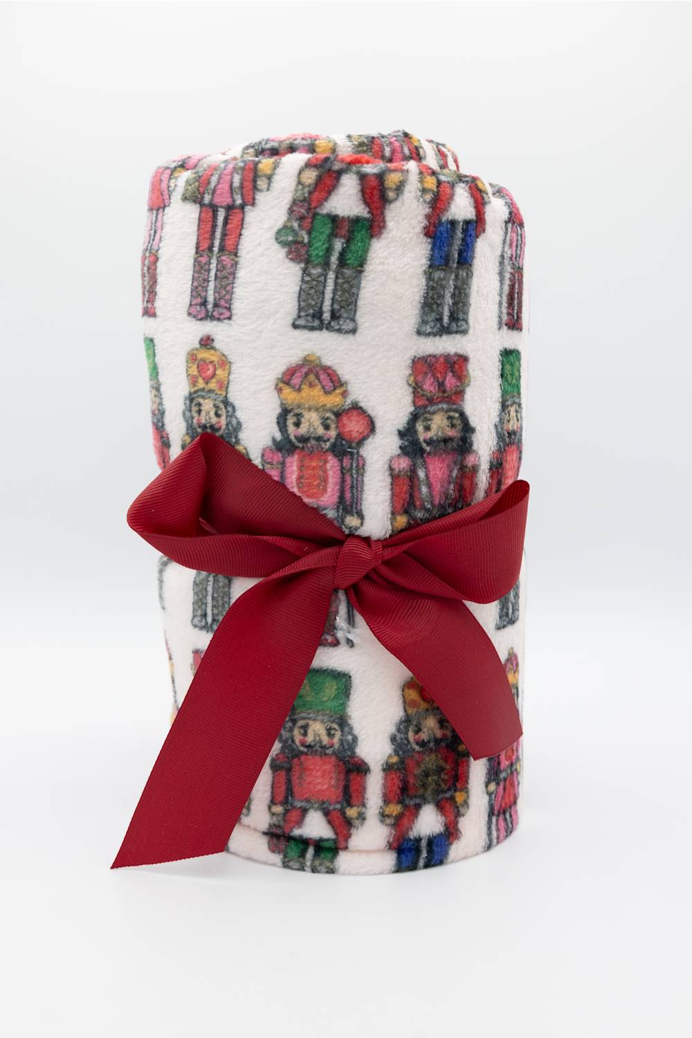 A rolled white fleece blanket featuring a colorful nutcracker pattern, tied with a large red satin bow, on a white background.