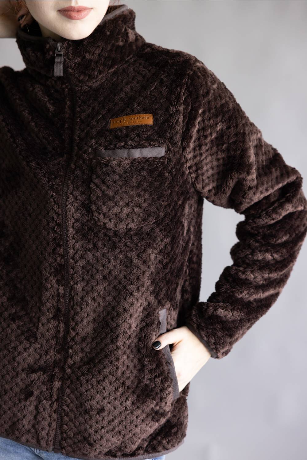 A close-up of a person wearing a dark brown textured fleece jacket with a high collar, front zipper, and a small tan accent patch on the chest pocket, set against a plain gray background.