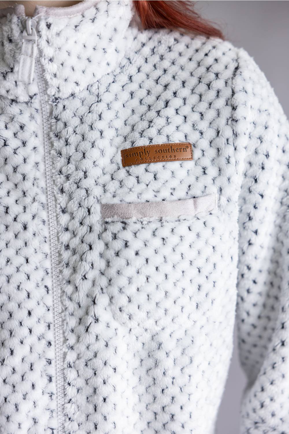 Close-up of a textured knit zip-up jacket in white and gray, featuring a small brown leather patch with the brand name 'Simply Southern' on the chest.