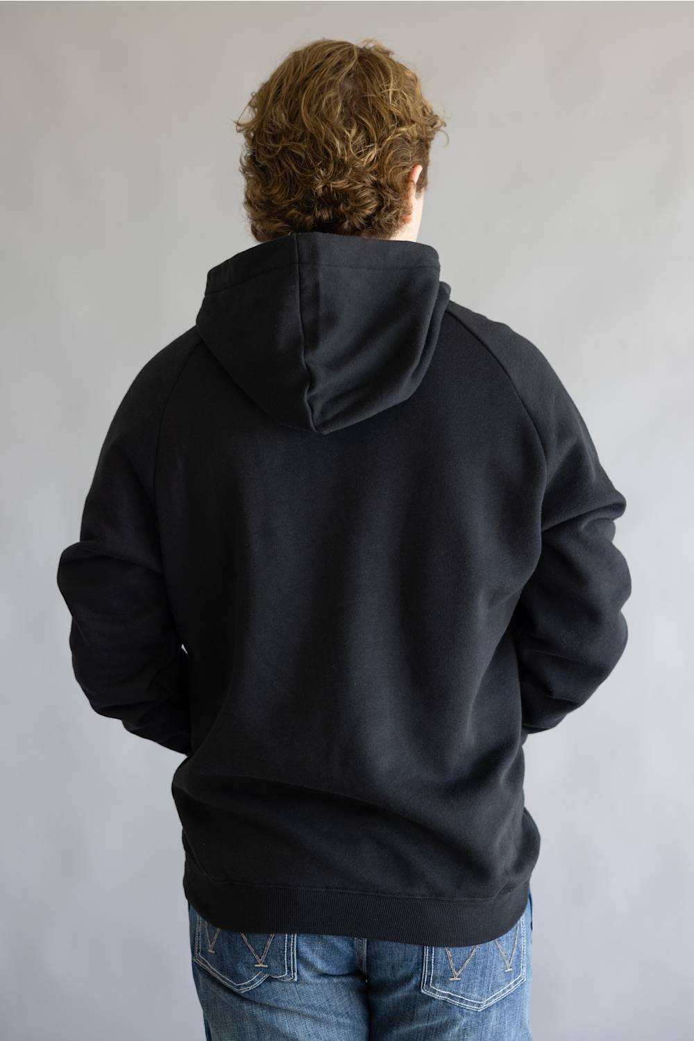A person viewed from behind wearing a black pullover hoodie and blue jeans against a plain light gray background.