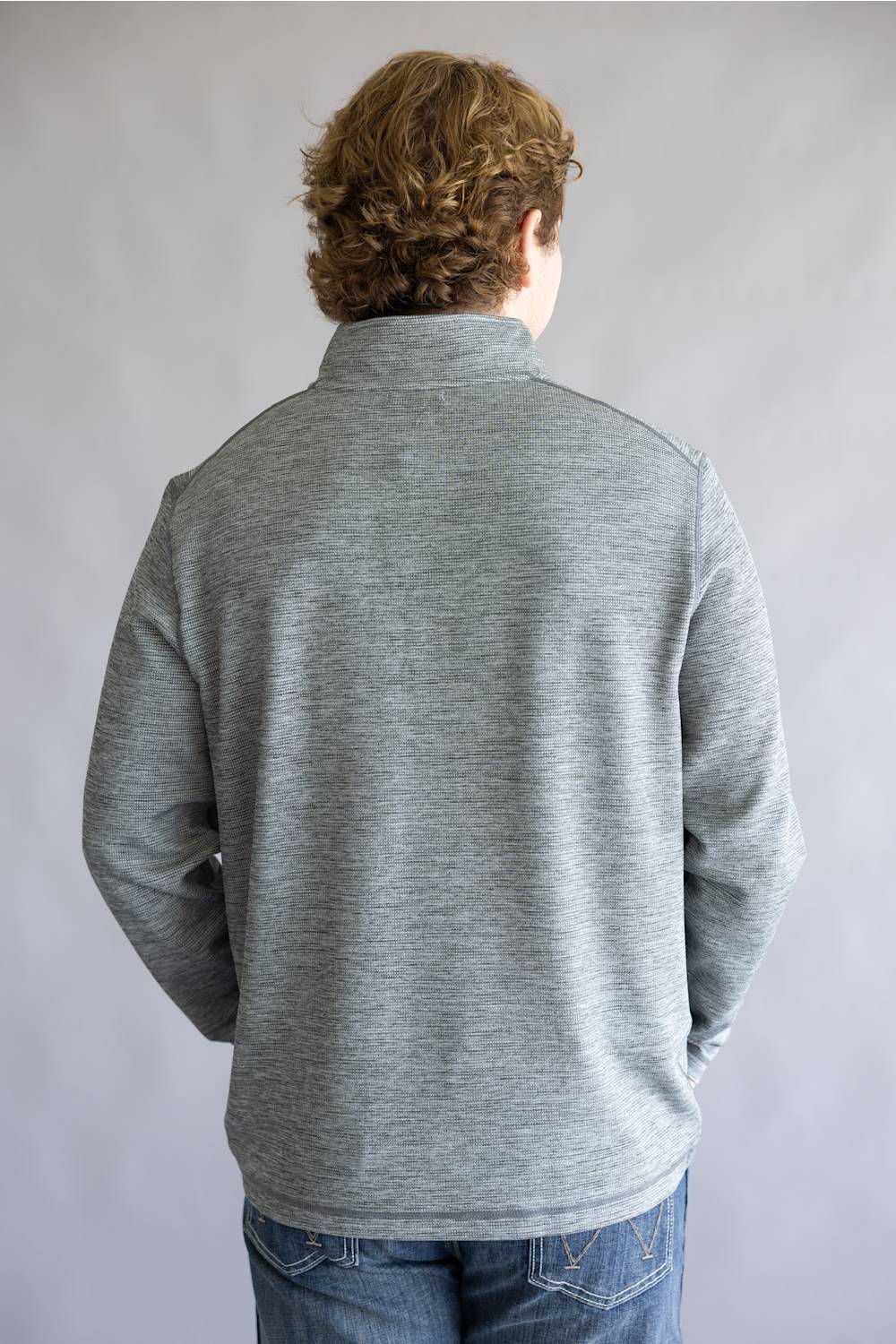A person viewed from the back wearing a heather gray quarter-zip pullover sweater and blue jeans, standing against a plain light gray studio background.