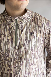 A close-up of a person wearing a camouflage-patterned quarter-zip pullover. The garment features an abstract wavy pattern in beige, brown, and muted purple-gray tones, with a dark olive green zipper and a small black embroidered deer logo on the left chest.