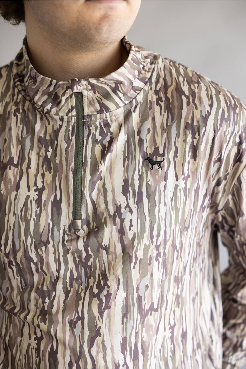 A close-up of a person wearing a camouflage-patterned quarter-zip pullover. The garment features an abstract wavy pattern in beige, brown, and muted purple-gray tones, with a dark olive green zipper and a small black embroidered deer logo on the left chest.