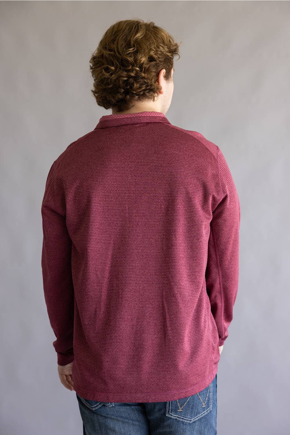 A person viewed from the back wearing a textured, long-sleeve collared knit top in a muted burgundy or maroon shade, paired with blue jeans, against a plain gray studio background.