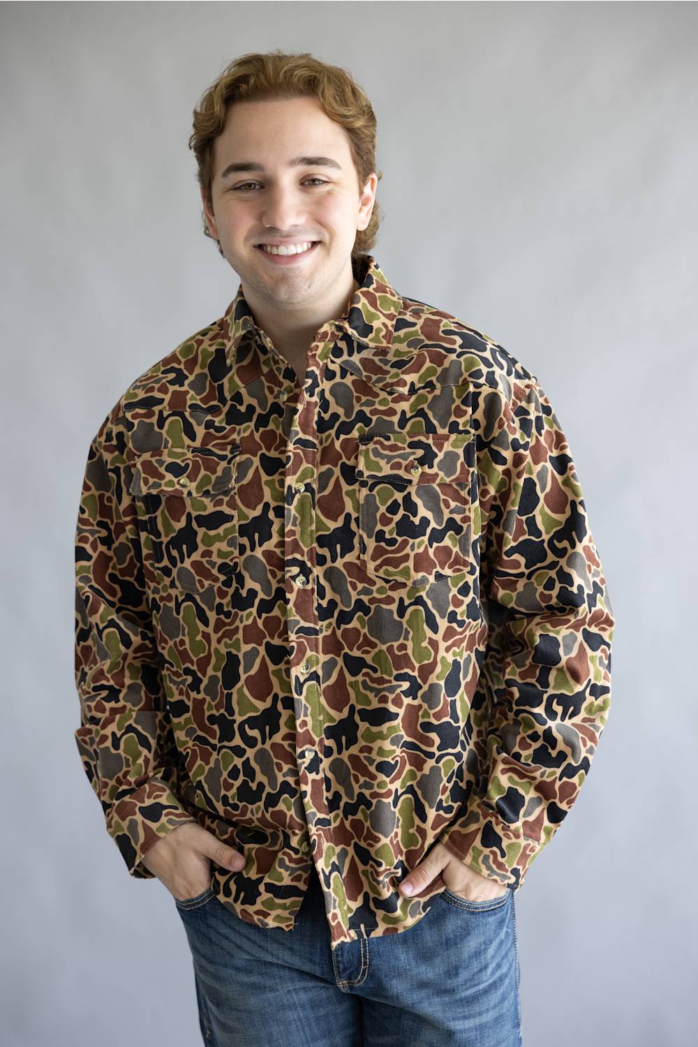 A smiling young man wearing a long-sleeve button-up shirt with an all-over camouflage pattern in brown, olive green, black, and tan, paired with blue jeans, standing against a neutral gray studio background.