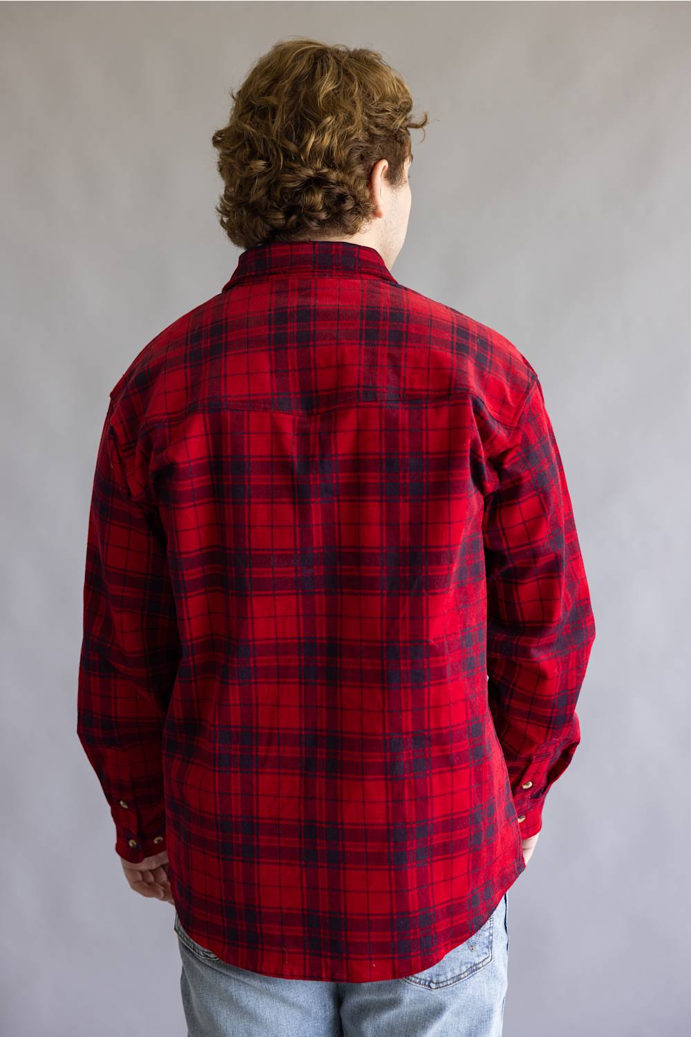 A person viewed from behind wearing a red and black plaid flannel shirt and light blue jeans against a plain gray studio background.