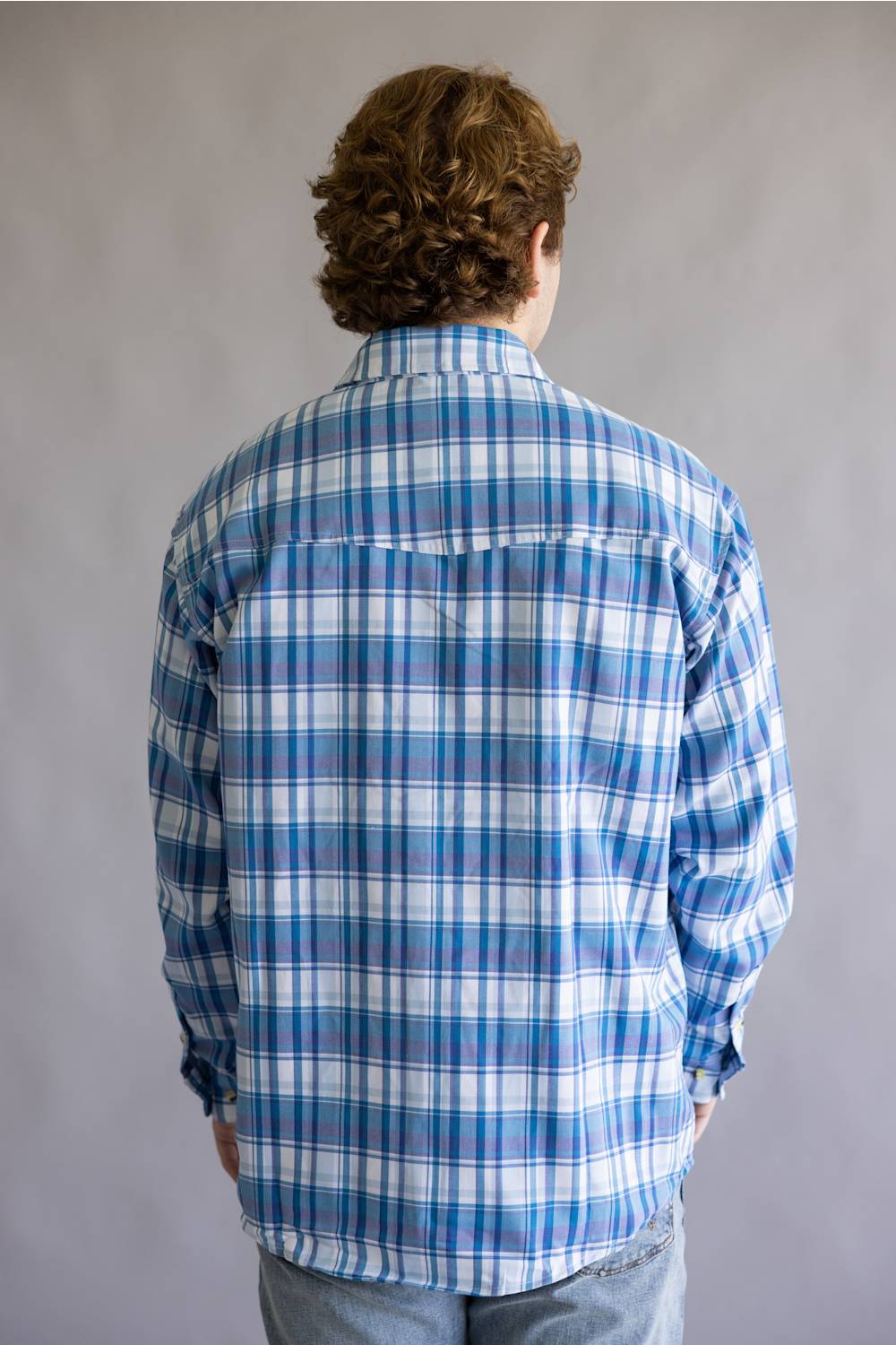 A person viewed from behind wearing a blue and white plaid button-up shirt and light blue jeans, standing against a plain gray studio background.