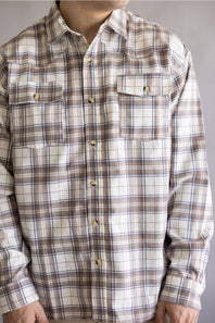 A man wearing a long-sleeve plaid button-up shirt in beige, brown, and white with gold-toned buttons, cropped from the neck down against a plain gray background.