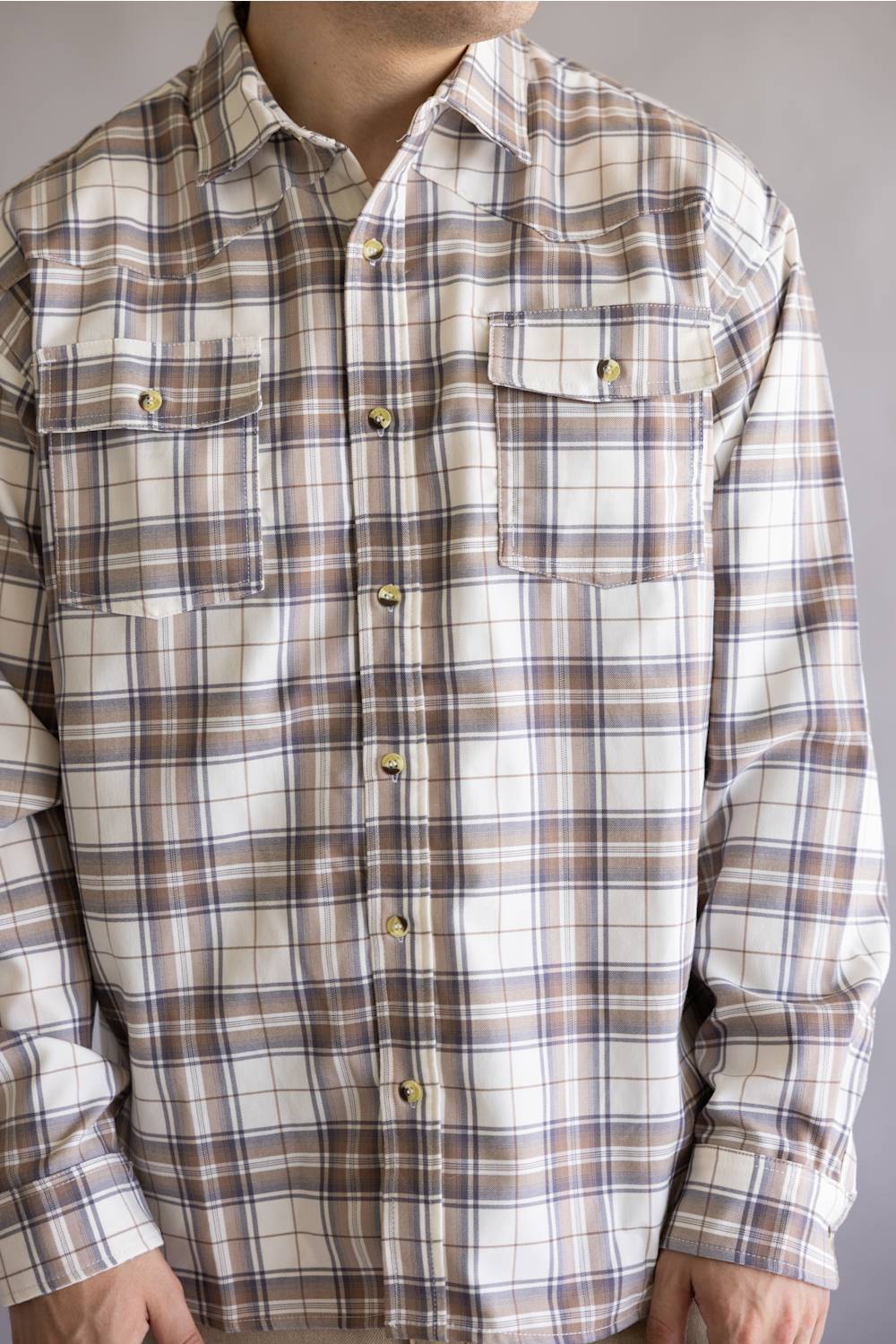 A man wearing a long-sleeve plaid button-up shirt in beige, brown, and white with gold-toned buttons, cropped from the neck down against a plain gray background.