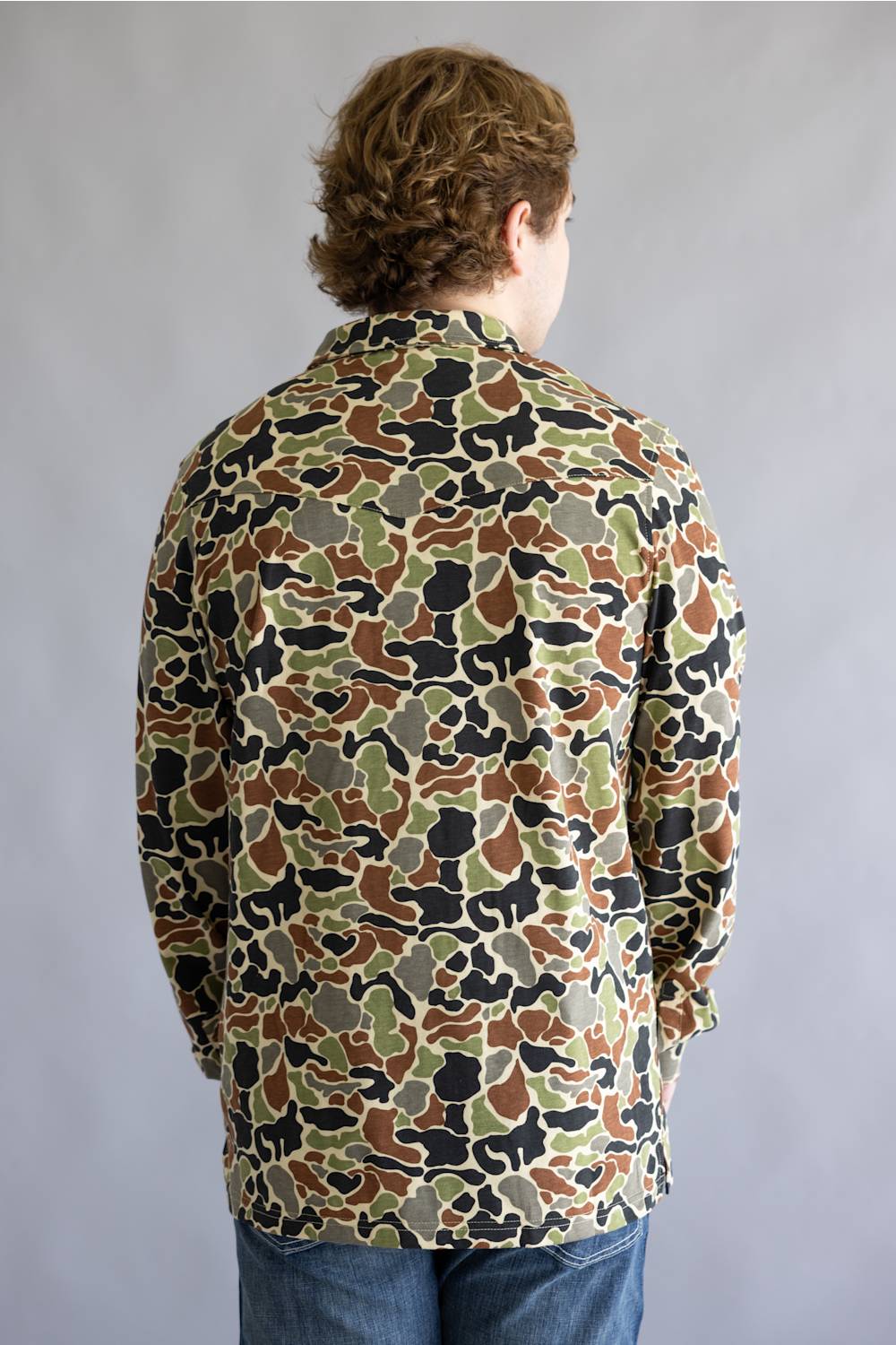 A man viewed from behind wearing a long-sleeve button-up shirt with an all-over camouflage pattern in olive green, brown, black, and beige, paired with blue jeans against a plain gray studio background.