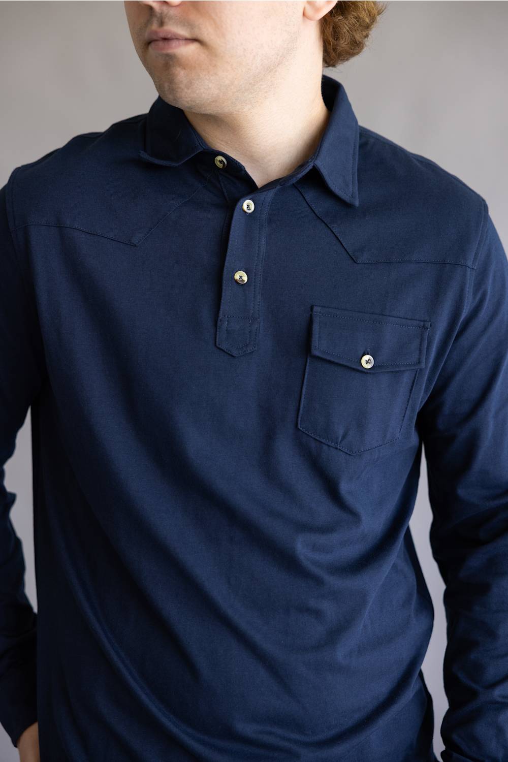 A man wearing a dark navy blue long-sleeve western-style shirt with a buttoned collar and chest pocket, shown from the chin down against a neutral gray background.