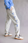 A person wearing light heather gray jogger sweatpants with blue bow embroidery down the side, paired with a blue sweatshirt and white sneakers with black stripes and gum soles, against a plain gray studio background.