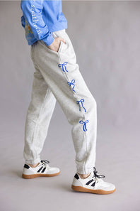 A person wearing light heather gray jogger sweatpants with blue bow embroidery down the side, paired with a blue sweatshirt and white sneakers with black stripes and gum soles, against a plain gray studio background.